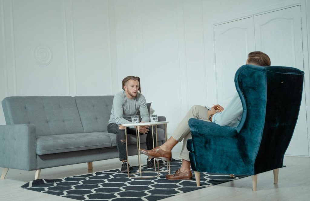 Two men engaged in a therapy session inside a modern office setting, discussing mental health.